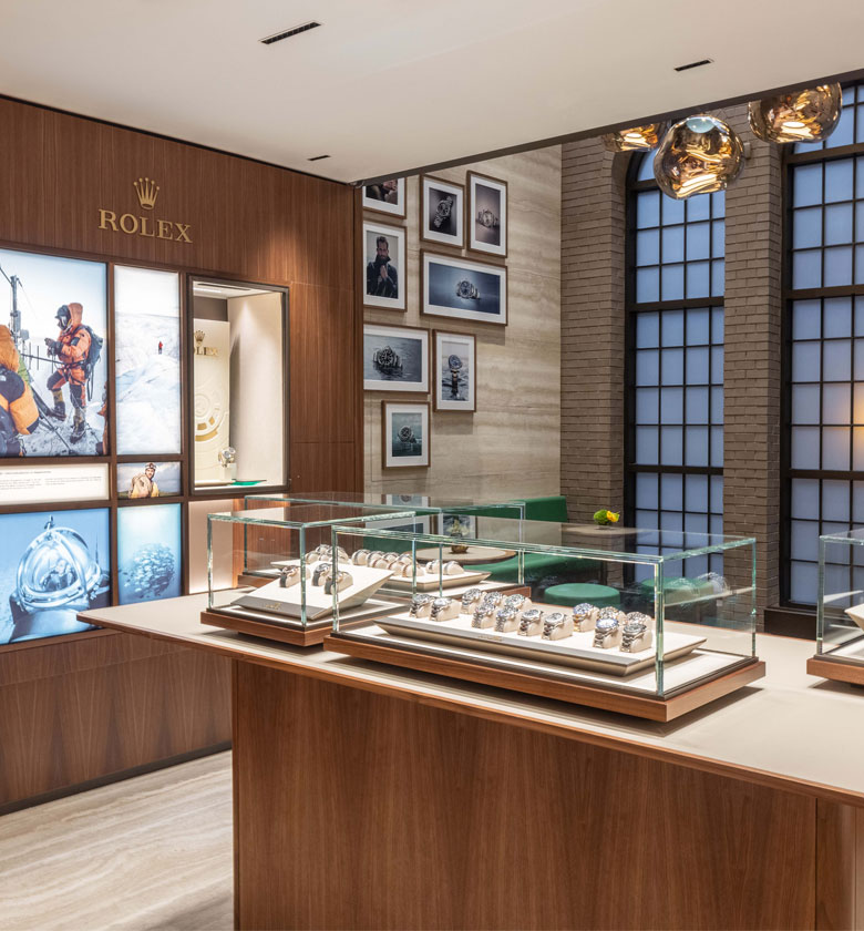 Interior of a Rolex store showing display cases filled with watches, a wall with framed photographs, and large windows with metal trim.