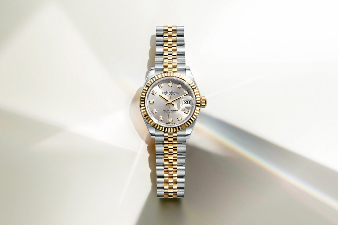 A gold watch with a metal link band, diamond-encrusted bezel, and a brown round dial with hour markers and hands. The background is a soft gradient of beige and white lights.