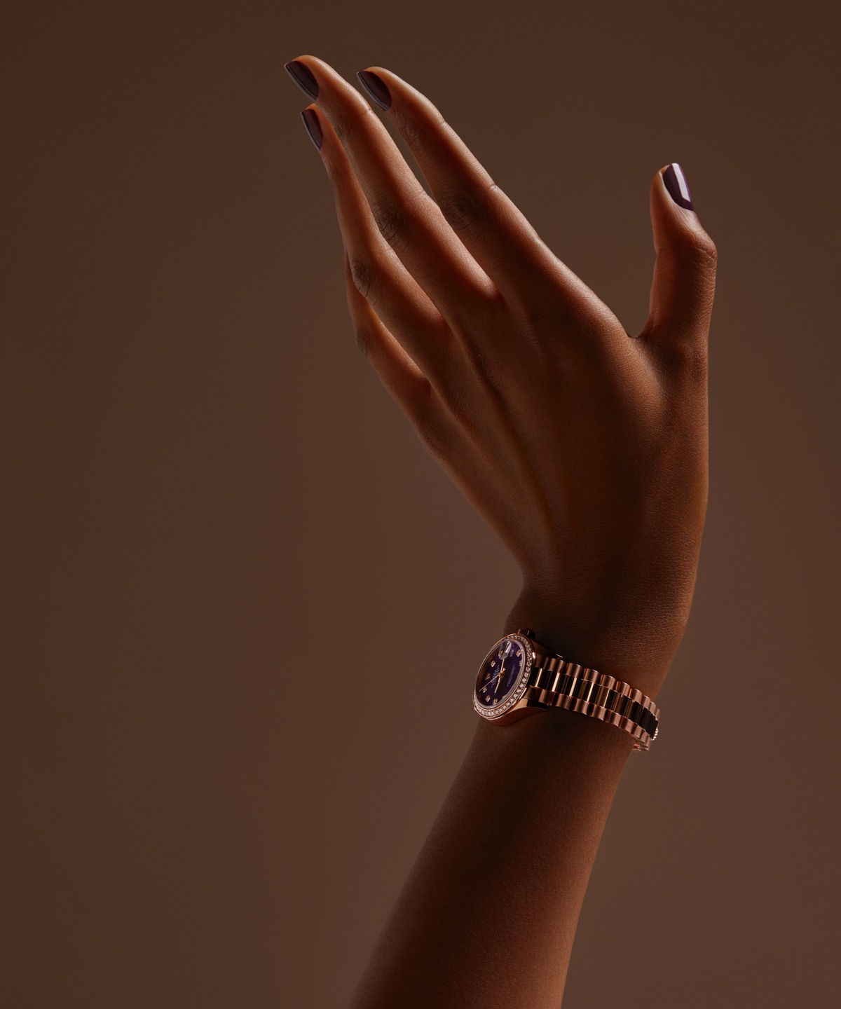 A hand with dark purple nail polish is raised against a brown background, wearing a gold watch.