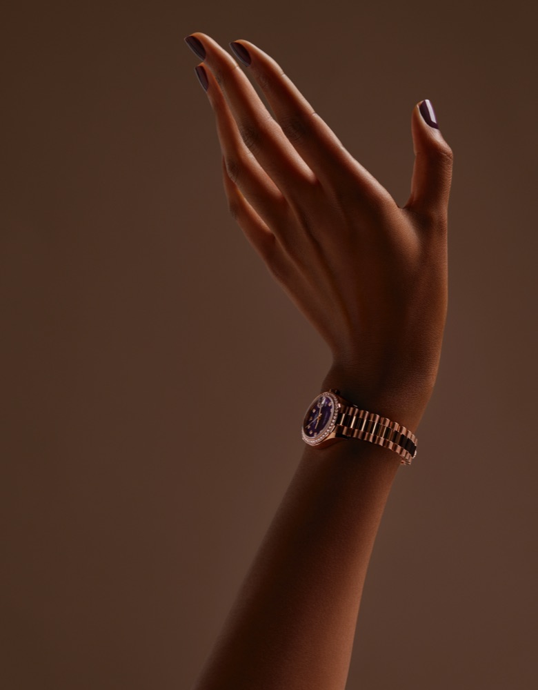 A hand with dark-painted nails is raised against a brown background, wearing a gold wristwatch.