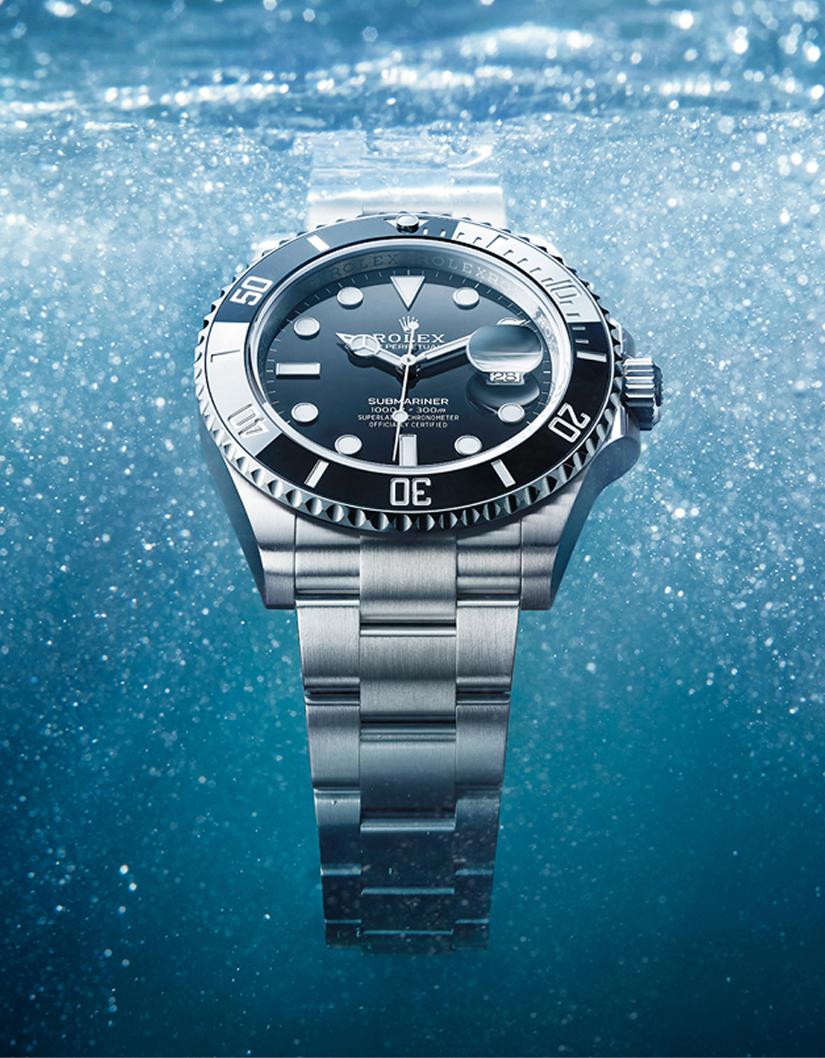 Close-up of a two-tone wristwatch with a blue face displaying the Rolex Submariner logo. The watch is underwater with air bubbles and a faint coral reef in the background.